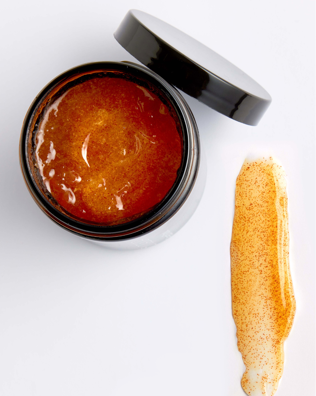 An open jar of BADESOFA Body Scrub on a plain white background, to the right shows a smear of a golden-yellow gel formula packed with fine, reddish-brown exfoliating particles