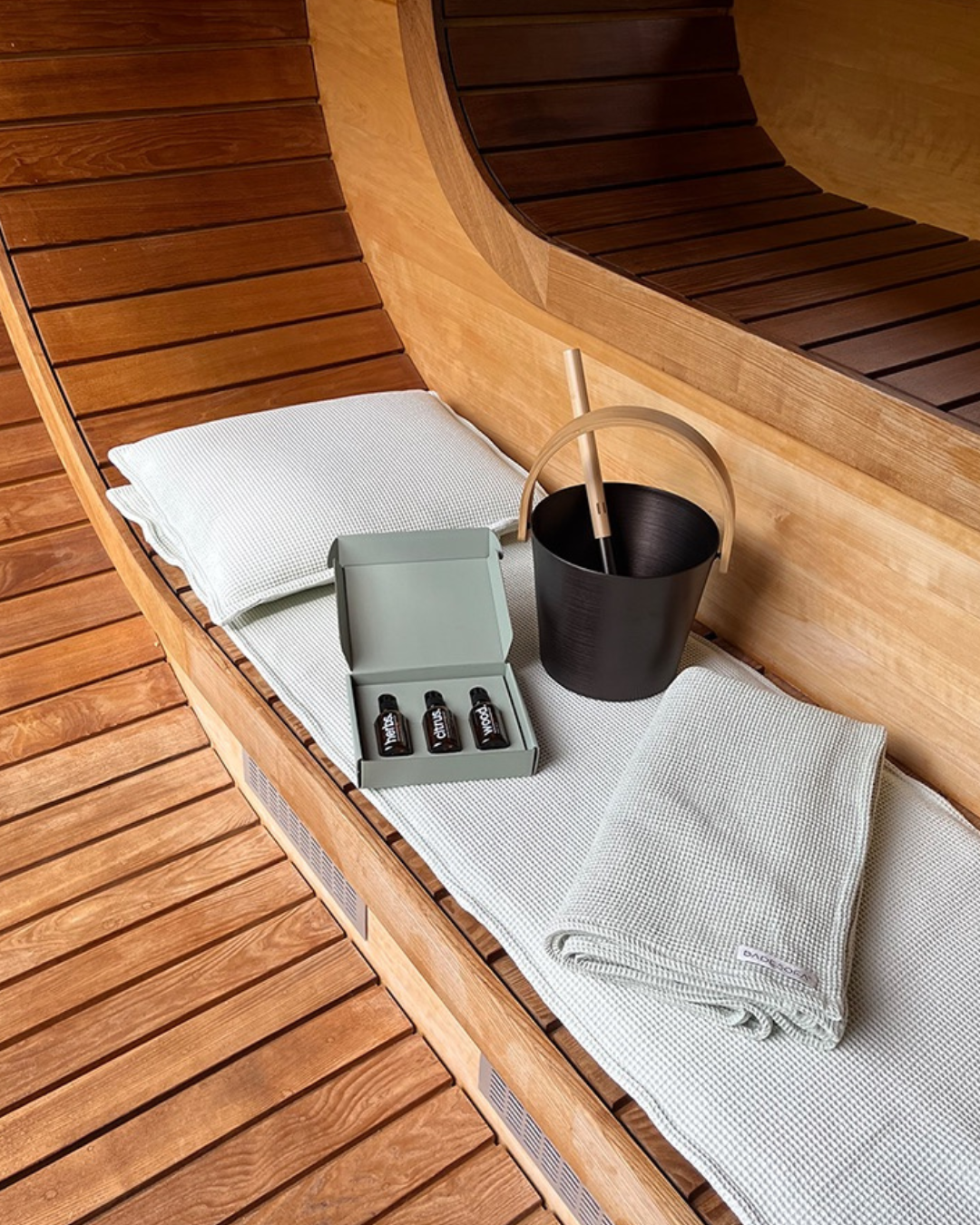 An image of the BADESOFA sauna set including a Black bucket with wooden handle, ladle, three essential oils in a box, a pillow, and a folded towel arranged on a wooden sauna bed