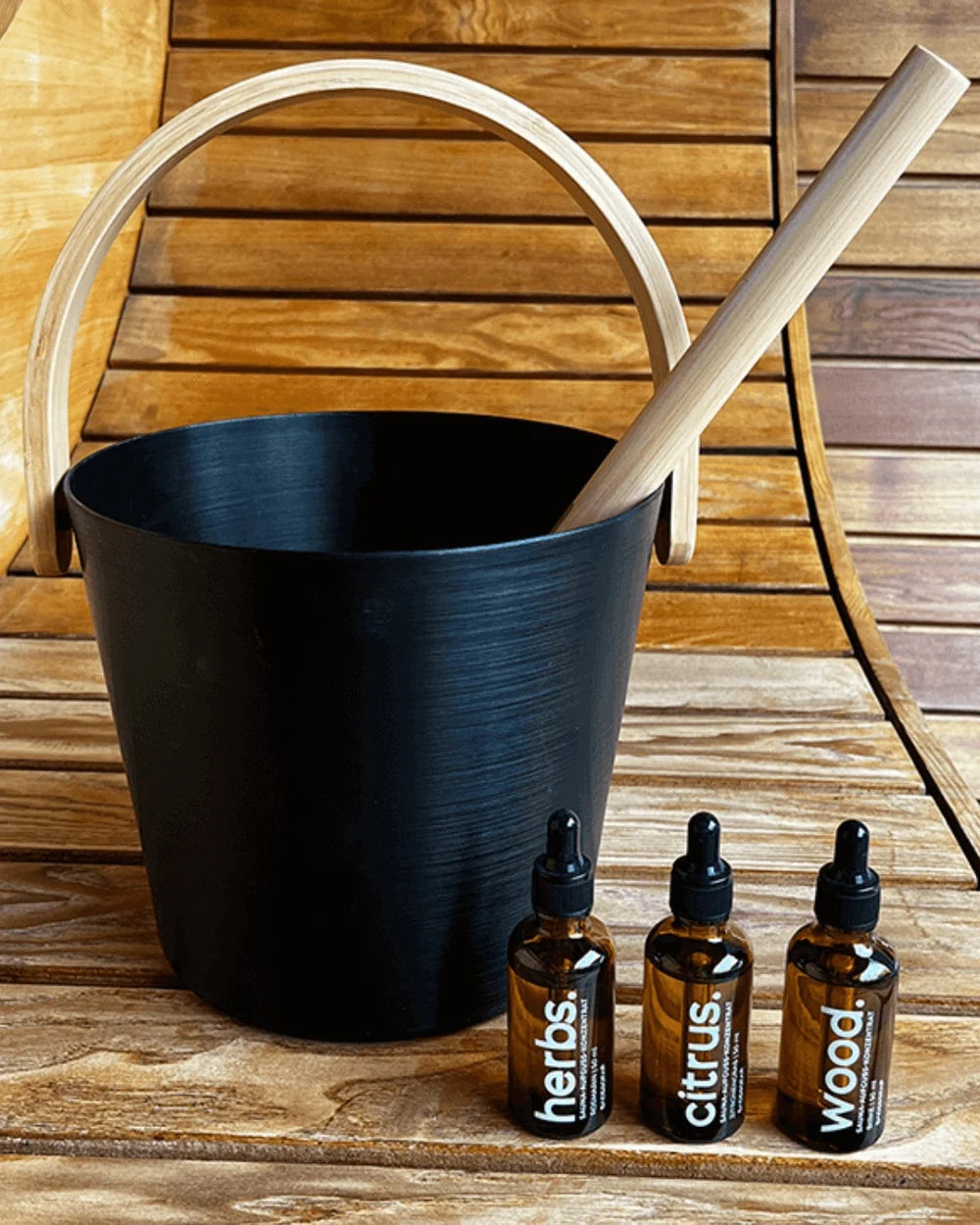 A close-up shot of a Black BADESOFA sauna bucket with a wooden handle and ladle on a wooden bench, with three essential oil bottles in front