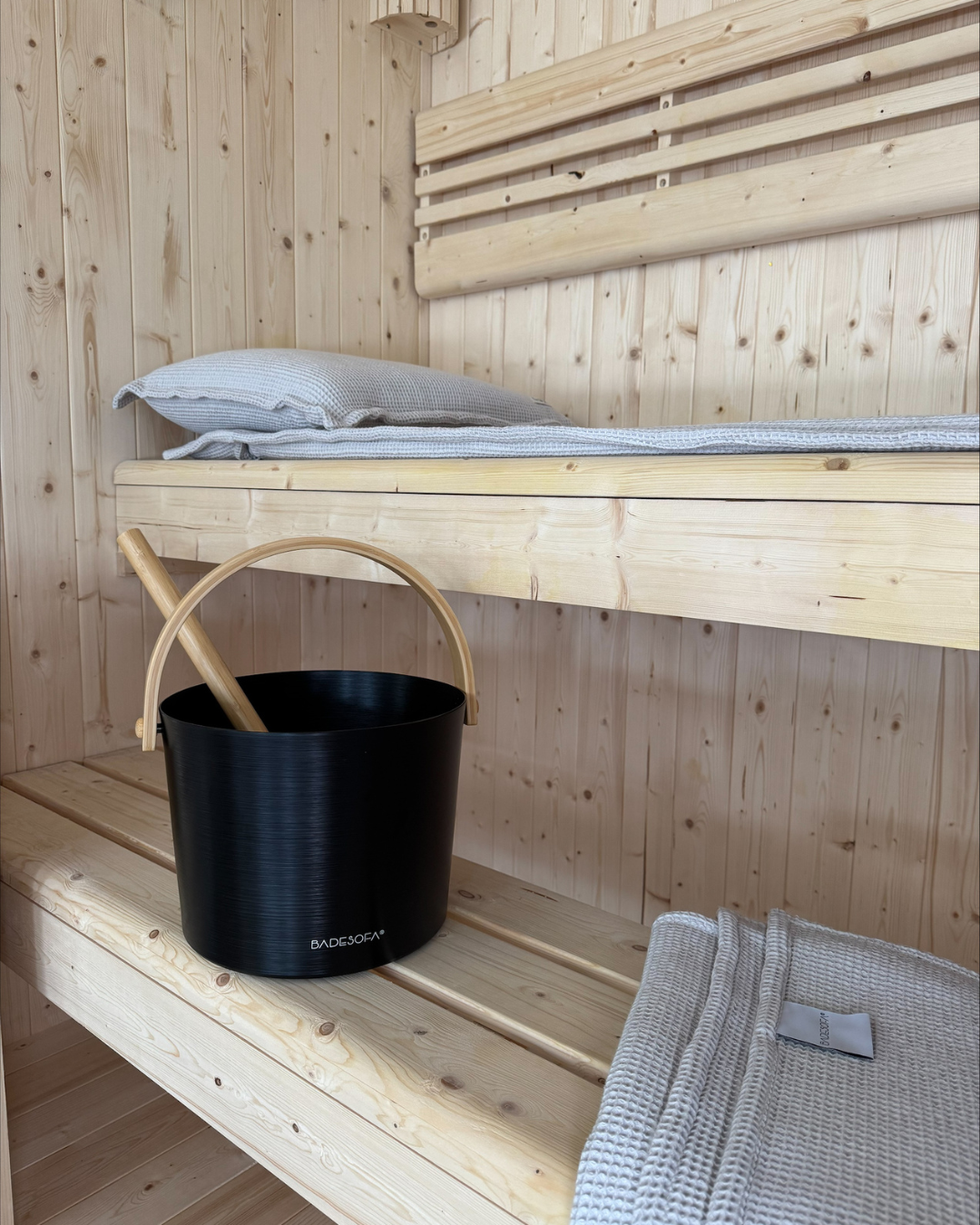 A photo of a Black BADESOFA sauna bucket with a light wooden handle and ladle sitting on a wooden sauna bench next to light Grey waffle-weave towels
