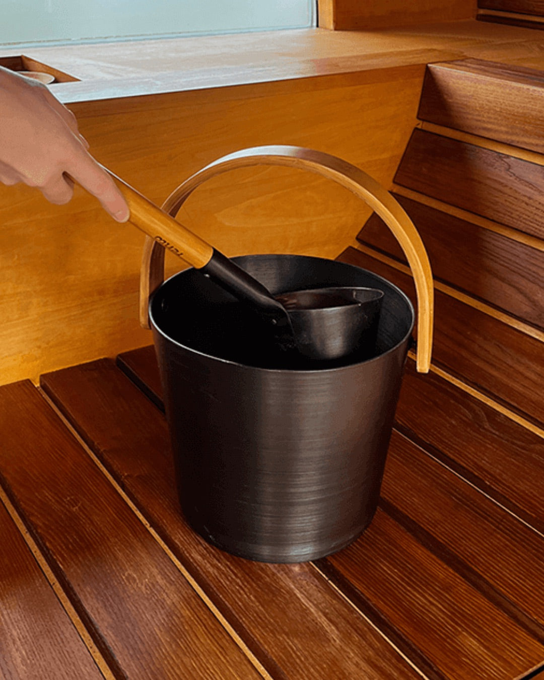 A photo of a hand reaching to place the Black BADESOFA wooden-handled ladle into the matching Black Aluminum sauna bucket on a wooden sauna bench